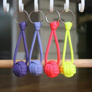 May include: Four colorful keychains hanging from a wooden bar. The keychains are purple, light blue, red, and neon yellow. Each keychain has a knotted ball at the bottom and a loop at the top for the key ring.
