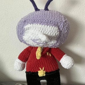 May include: A handmade plush toy in the shape of a cartoon character. The toy has a lavender head with two purple antennae, a white face, a red shirt, black pants, and yellow accents. The hands and feet are white and yellow.