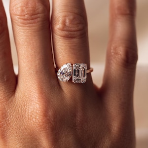 Può includere: Un anello d'oro con due diamanti: una pietra a forma di pera e una pietra taglio smeraldo. L'anello è indossato su un dito, evidenziando la brillantezza dei diamanti chiari e sfaccettati. La fascia è in oro sottile.