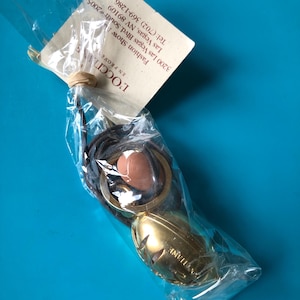 May include: A gold-coloured metal charm with a decorative design hangs from a brown cord. The charm is packaged in a clear plastic bag with a white tag that reads "L'OCCITANE Fashion Show 3200 Las Vegas Blvd. South 2005 Las Vegas, NV 89109 Tel: (702) 369-1286".