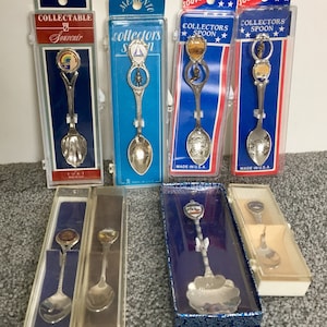 May include: A collection of eight silver souvenir spoons in individual packaging. The spoons feature various designs, including a lighthouse, a ship, and a fort. The packaging is blue, red, and white with the words "Collector's Spoon" and "Made in U.S.A."