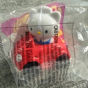 May include: A white Hello Kitty figurine in a clear plastic bag. The figurine has a red bow and blue outfit. The packaging includes text in English and French, with a warning about small parts.
