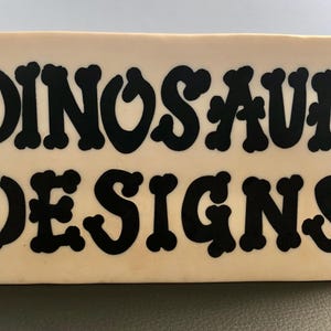 May include: A white rectangular sign with black lettering that reads "Dinosaur Designs".