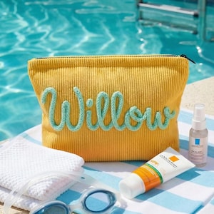 Pode incluir: Bolsa de cosm&eacute;ticos de veludo cotel&ecirc; amarelo mostarda com o nome "Willow" em escrita azul clara. A bolsa est&aacute; ao lado de uma toalha branca, protetor solar, n&eacute;voa facial e &oacute;culos de nata&ccedil;&atilde;o azuis. Os itens est&atilde;o em uma toalha listrada perto de uma piscina.