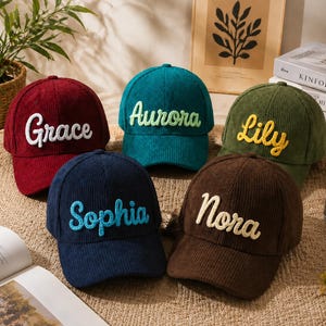 May include: Five corduroy baseball caps in burgundy, teal, olive green, navy blue, and brown. Each cap features embroidered names in contrasting colours: Grace in white, Aurora in light green, Lily in yellow, Sophia in light blue, and Nora in cream.
