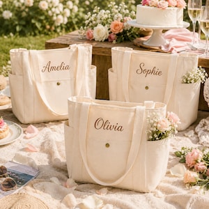 Peut inclure: Trois sacs fourre-tout en toile blanc cassé dotés de deux poches latérales, d'une poche avant avec un bouton-pression en laiton et de prénoms brodés en écriture brune. Les sacs sont nommés Amelia, Sophie et Olivia, avec deux anses solides.