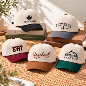 Puede incluir: Cinco gorras de béisbol de pana con coronas color crema y viseras en contraste. Las gorras presentan diseños bordados: una hoja de arce negra con "TRUE NORTH", "COZY CLUB" con árboles, "EH?" en granate, "Weekend" en marrón y "LAKE LIFE" con montañas.