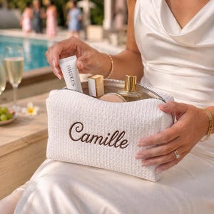 May include: A white waffle-textured fabric toiletry bag with a zip closure, featuring the name Camille embroidered in brown script on the front. The bag is open and contains a white tube labelled HIISEES, a gold-capped bottle, and a compact.