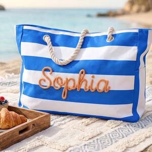 May include: A blue and white striped beach bag with the name "Sophia" in orange script. The bag has rope handles. A tray with a croissant and a bowl of berries sits in front of the bag on a beach. The sea is in the background.