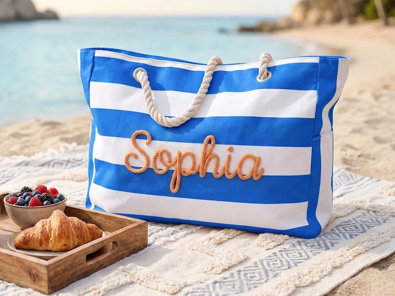 May include: A blue and white striped beach bag with the name "Sophia" in orange script. The bag has rope handles. A tray with a croissant and a bowl of berries sits in front of the bag on a beach. The sea is in the background.