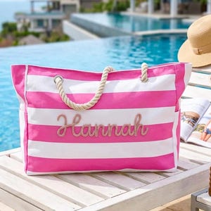 May include: A pink and white striped beach bag with rope handles. The name "Hannah" is embroidered on the front. The bag is on a wooden surface with a hat, sunglasses, and a magazine.