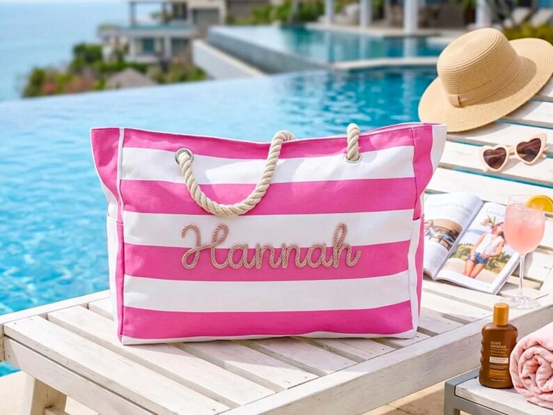 May include: A pink and white striped beach bag with rope handles. The name "Hannah" is embroidered on the front. The bag is on a wooden surface with a hat, sunglasses, and a magazine.