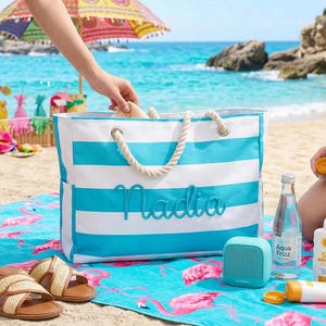May include: A beach scene with a blue and white striped tote bag embroidered with "Nadia". The bag has rope handles and is next to a flamingo-patterned beach towel. Other items include sandals, a speaker, sunscreen, and a water bottle.