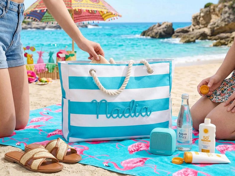 May include: A beach scene with a blue and white striped tote bag embroidered with "Nadia". The bag has rope handles and is next to a flamingo-patterned beach towel. Other items include sandals, a speaker, sunscreen, and a water bottle.