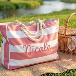 May include: A pink and white striped tote bag embroidered with the name "Nicole". The bag has rope handles and is placed next to a picnic basket, wine glass, and a bottle of ros&eacute; wine on a blanket.
