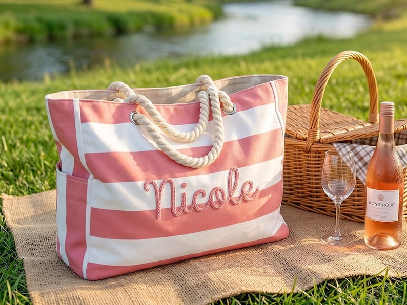 May include: A pink and white striped tote bag embroidered with the name "Nicole". The bag has rope handles and is placed next to a picnic basket, wine glass, and a bottle of ros&eacute; wine on a blanket.