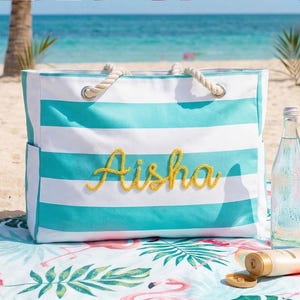 May include: A striped beach bag in turquoise and white, with the name "Aisha" embroidered in yellow. The bag has rope handles and is on a beach towel with a flamingo design. A water bottle and sunscreen are also visible.