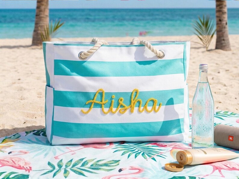 May include: A striped beach bag in turquoise and white, with the name "Aisha" embroidered in yellow. The bag has rope handles and is on a beach towel with a flamingo design. A water bottle and sunscreen are also visible.