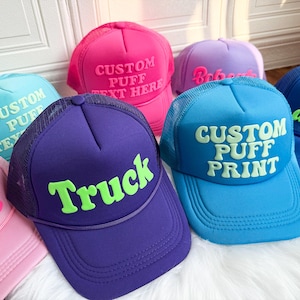 以下が含まれることがあります： メッシュバック付きのカラフルなトラッカーハットのコレクション。帽子は、青、ピンク、紫など、さまざまな色があり、「Truck」、「Custom Puff Print」などのカスタムテキストが白と緑で表示されています。