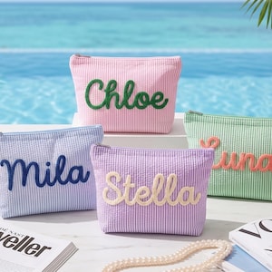 Peut inclure: Quatre trousses de maquillage en seersucker rose, bleu, violet et vert, chacune avec un nom différent brodé dans une couleur contrastante. Les sacs sont présentés sur une surface blanche avec une piscine en arrière-plan.