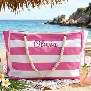 Könnte beinhalten: Eine große Strandtasche mit rosa und weißen Streifen und Seilgriffen. Der Name "Olivia" ist auf der Vorderseite gestickt. Die Tasche befindet sich an einem Strand mit einem Getränk, einer Sonnenbrille und Sonnencreme.
