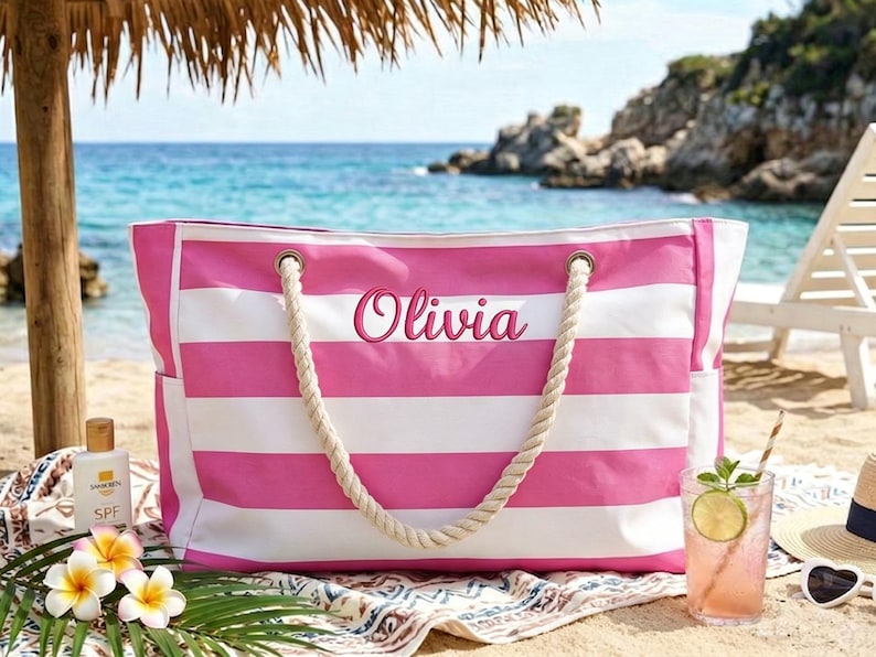 May include: A pink and white striped beach tote bag with rope handles. The name "Olivia" is embroidered on the front. The bag is on a beach with a drink, sunglasses, and sunscreen.