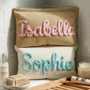 Pode incluir: Duas bolsas de cosm&eacute;ticos de veludo cotel&ecirc;, uma bege com "Isabella" em fio rosa, a outra creme com "Sophie" em fio azul. Produtos de cuidados com a pele e uma barra de sab&atilde;o s&atilde;o dispostos ao redor das bolsas.