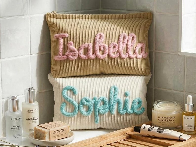 Pode incluir: Duas bolsas de cosm&eacute;ticos de veludo cotel&ecirc;, uma bege com "Isabella" em fio rosa, a outra creme com "Sophie" em fio azul. Produtos de cuidados com a pele e uma barra de sab&atilde;o s&atilde;o dispostos ao redor das bolsas.