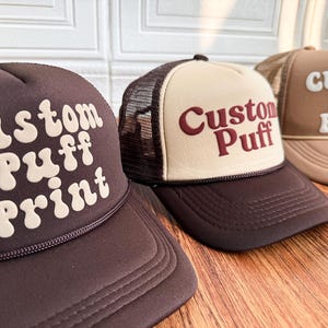 May include: Three trucker hats with the text "Custom Puff Print". The hats are brown, beige and tan with mesh backs and curved brims. The hats are displayed on a wooden surface.