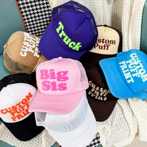 May include: A selection of trucker caps in diverse colours, including blue, pink, brown, and white. Each cap displays text like "Truck", "Big Sis", and "Custom Puff Print". The caps have mesh backs and curved peaks.