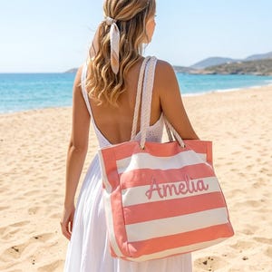 May include: A coral and white striped tote bag with the name "Amelia" in cursive script. The bag has rope handles and is being carried on a beach. The background features a blue ocean and sandy beach.