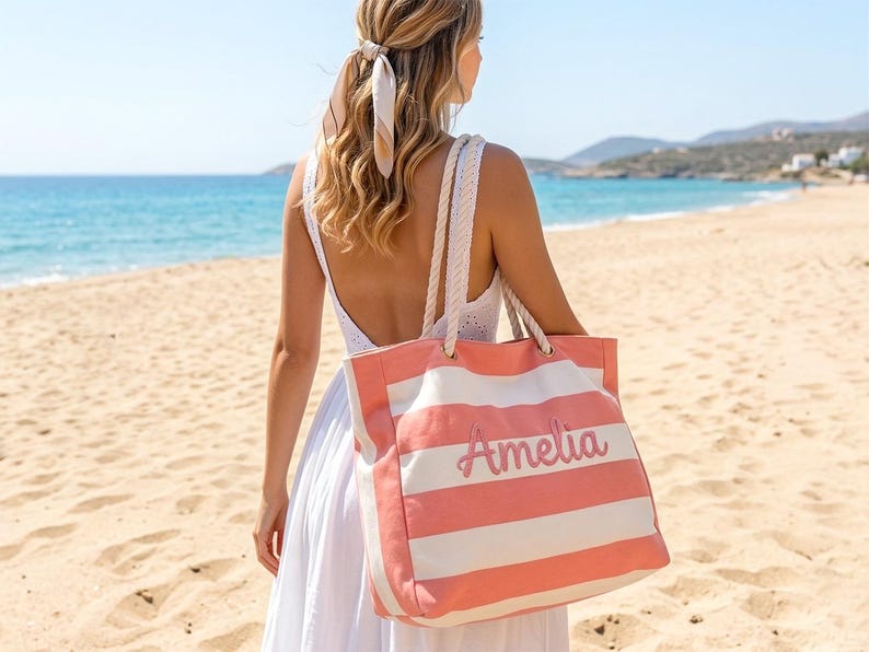 May include: A coral and white striped tote bag with the name "Amelia" in cursive script. The bag has rope handles and is being carried on a beach. The background features a blue ocean and sandy beach.