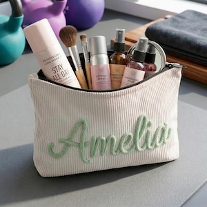 Pode incluir: Uma bolsa de maquilhagem de veludo cotel&ecirc; creme com o nome "Amelia" bordado a verde. A bolsa cont&eacute;m pinc&eacute;is de maquilhagem, spray fixador e outros produtos de beleza. O texto "STAY ALL DAY" &eacute; vis&iacute;vel num produto. A bolsa est&aacute; sobre um tapete cinzento.