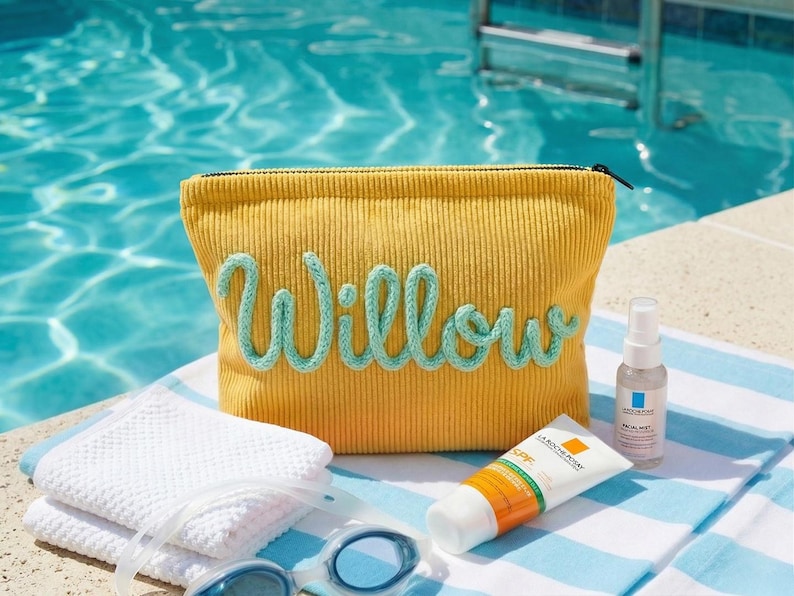 Pode incluir: Bolsa de cosm&eacute;ticos de veludo cotel&ecirc; amarelo mostarda com o nome "Willow" em escrita azul clara. A bolsa est&aacute; ao lado de uma toalha branca, protetor solar, n&eacute;voa facial e &oacute;culos de nata&ccedil;&atilde;o azuis. Os itens est&atilde;o em uma toalha listrada perto de uma piscina.