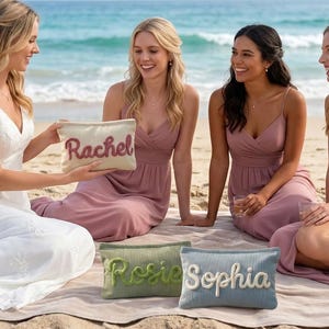 Pode incluir: Quatro mulheres numa praia, uma com um vestido branco, outras com vestidos rosa, reunidas em torno de n&eacute;cessaires personalizados. As bolsas s&atilde;o bege, verdes e azuis, com nomes bordados em v&aacute;rias cores. O cen&aacute;rio &eacute; uma praia com areia e ondas.