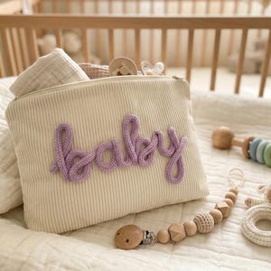 Pode incluir: Uma bolsa de beb&ecirc; de veludo cotel&ecirc; creme com a palavra "baby" em escrita lavanda. A bolsa &eacute; preenchida com artigos para beb&ecirc;s e est&aacute; ao lado de uma chocalho de madeira, um prendedor de chupeta e um chocalho de cordeiro de croch&ecirc;. Os itens est&atilde;o em um cobertor branco acolchoado.