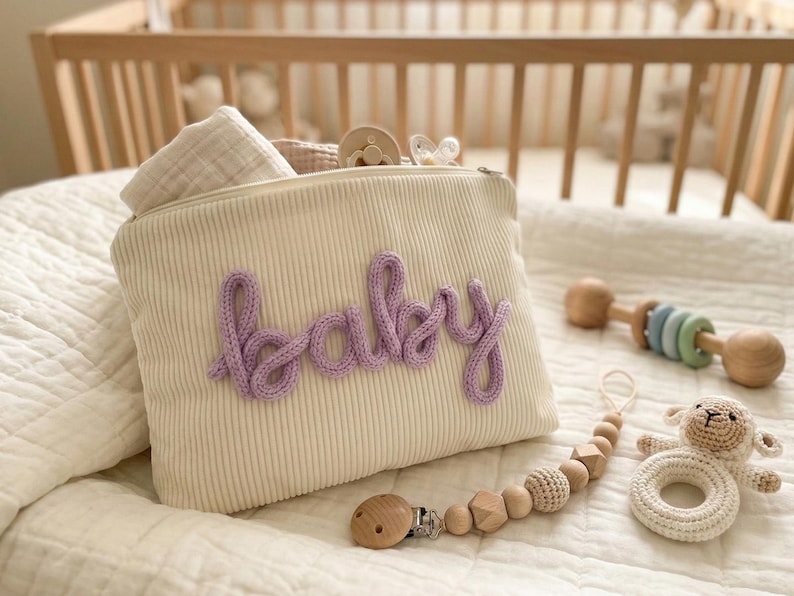 Pode incluir: Uma bolsa de beb&ecirc; de veludo cotel&ecirc; creme com a palavra "baby" em escrita lavanda. A bolsa &eacute; preenchida com artigos para beb&ecirc;s e est&aacute; ao lado de uma chocalho de madeira, um prendedor de chupeta e um chocalho de cordeiro de croch&ecirc;. Os itens est&atilde;o em um cobertor branco acolchoado.