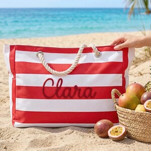 May include: A red and white striped beach tote bag with the name "Clara" embroidered in red script. The bag has rope handles and sits on a sandy beach next to a basket of tropical fruit, including mangoes, passion fruit, and a pineapple.