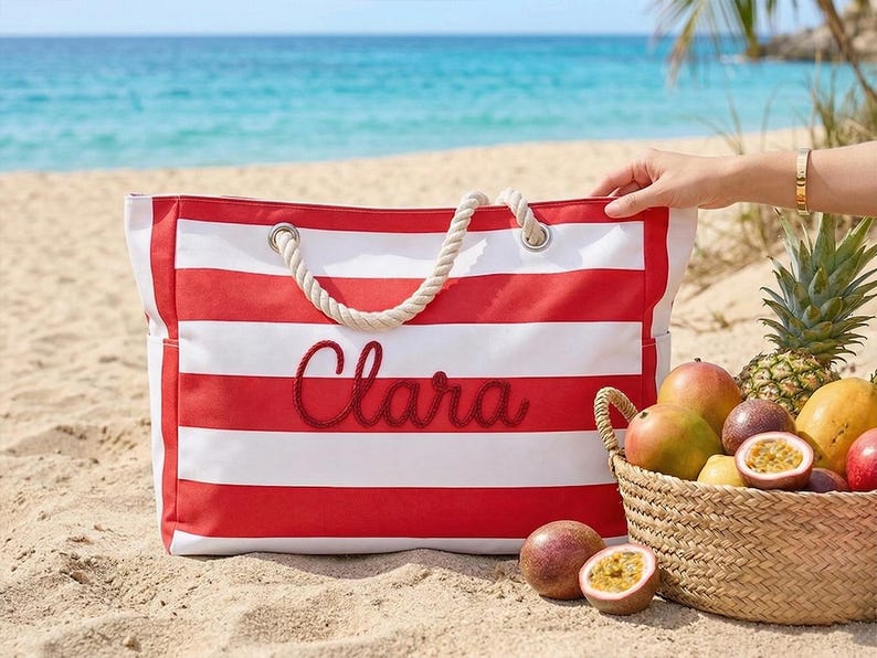 May include: A red and white striped beach tote bag with the name "Clara" embroidered in red script. The bag has rope handles and sits on a sandy beach next to a basket of tropical fruit, including mangoes, passion fruit, and a pineapple.