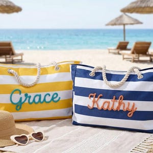 May include: Two striped beach tote bags with rope handles. One is yellow and white with "Grace" in teal script, the other is blue and white with "Kathy" in coral script. A straw hat and heart-shaped sunglasses are in the foreground on the beach.
