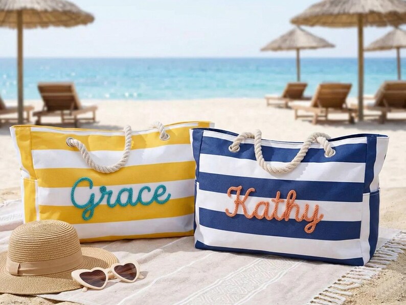 May include: Two striped beach tote bags with rope handles. One is yellow and white with "Grace" in teal script, the other is blue and white with "Kathy" in coral script. A straw hat and heart-shaped sunglasses are in the foreground on the beach.