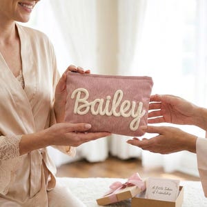 Pode incluir: Uma bolsa de cosm&eacute;ticos de veludo cotel&ecirc; rosa com o nome "Bailey" em escrita branca. A bolsa est&aacute; sendo segurada por duas pessoas. Uma pequena caixa de presente com um la&ccedil;o rosa est&aacute; em primeiro plano.