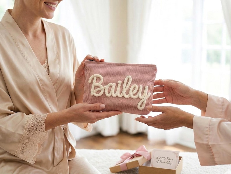 Pode incluir: Uma bolsa de cosm&eacute;ticos de veludo cotel&ecirc; rosa com o nome "Bailey" em escrita branca. A bolsa est&aacute; sendo segurada por duas pessoas. Uma pequena caixa de presente com um la&ccedil;o rosa est&aacute; em primeiro plano.