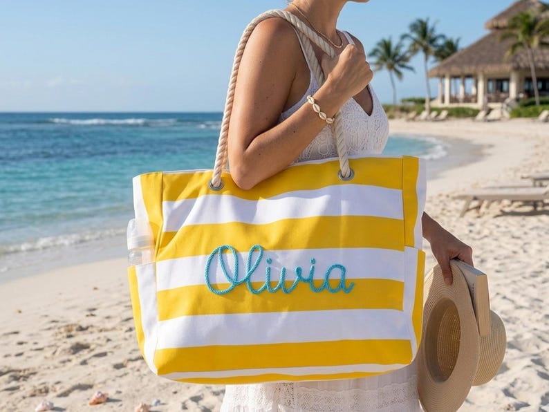 May include: A yellow and white striped tote bag with the name "Olivia" embroidered in turquoise. The bag has rope handles and side pockets. A straw hat is held in the hand. The background shows a beach and ocean.