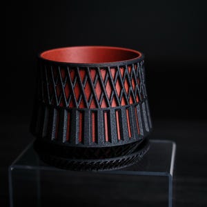 May include: A black and red decorative planter with a geometric design. The planter has a cylindrical shape with a textured exterior and a contrasting red interior. It sits on a matching saucer.