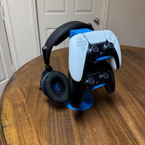 May include: A blue and black gaming headset and two game controllers rest on a blue stand. One controller is white, the other is black. The stand is on a wooden table.