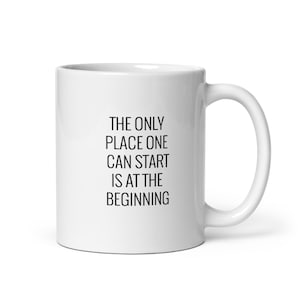 May include: White ceramic mug with a curved handle. The mug features the text "THE ONLY PLACE ONE CAN START IS AT THE BEGINNING" in black, centered on the front. The mug is designed for hot beverages.