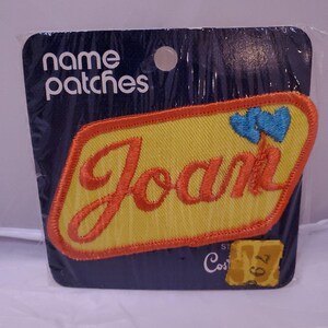 May include: A yellow iron-on name patch with the name "Joan" in red lettering and two blue hearts. The patch is packaged on a blue card with the text "name patches" in white lettering.
