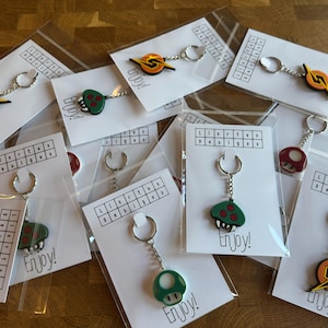 May include: Keychains with various designs, including a mushroom, a lightning bolt, and a red mushroom. Each keychain is attached to a silver keyring and packaged on a white card with the word "Enjoy!" printed on it. The keychains are in clear plastic bags.