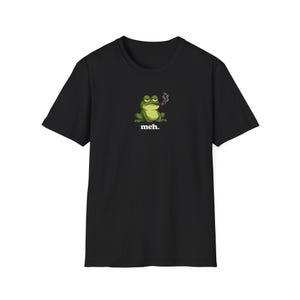 May include: A black short-sleeved t-shirt featuring a graphic of a green frog with a bored expression, smoking a cigar with a small trail of smoke rising. Below the frog, the word "meh." is printed in a bold, white, lowercase sans-serif font.
